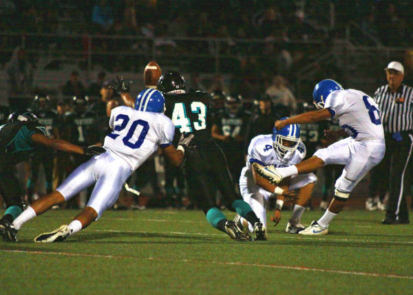 A high school football kicker in a white and blue uniform attempting a field goal during a night game.