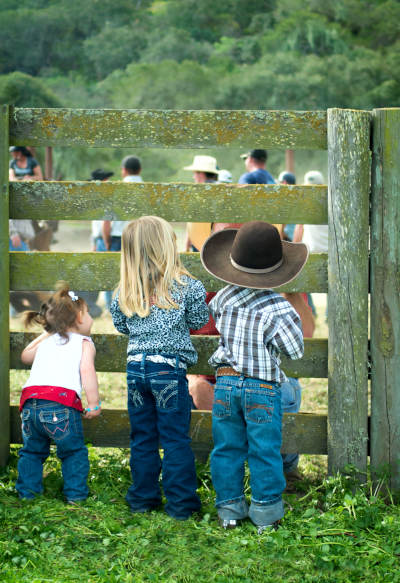 Three young children in cowboy hats and denim watching a rodeo event through a rustic wooden fence.
