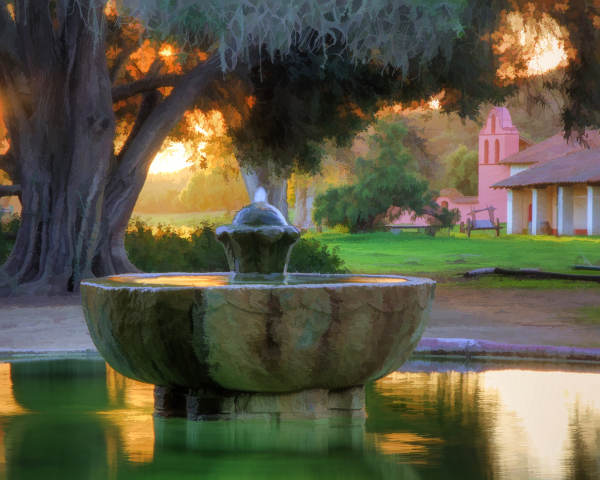 A stone water fountain in the courtyard of a Spanish mission at sunset, surrounded by green grass and large oak trees.
