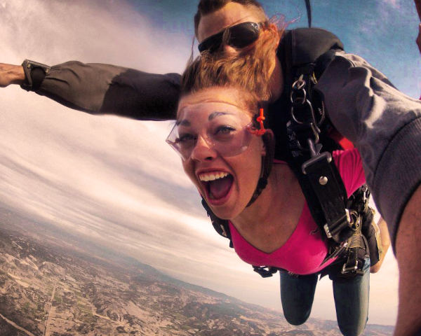 A high-angle selfie of a woman smiling and screaming with excitement during a tandem skydiving jump over a landscape.