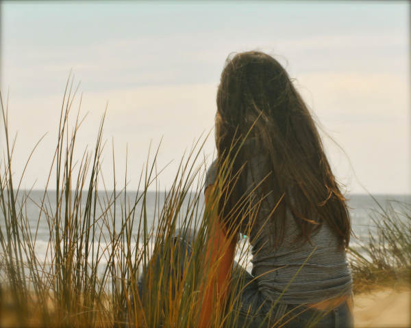 A girl with long hair sitting among tall beach grass, looking out toward the ocean on a cloudy day.